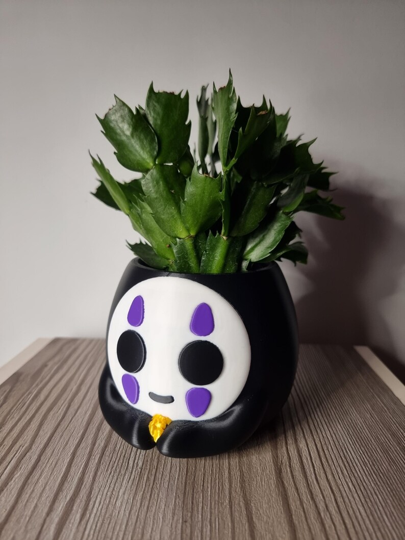 May include: A black and white planter shaped like a character with a white face, black circular eyes, and purple teardrop markings above and below the eyes. The character holds a small yellow object. A green succulent plant grows from the top.