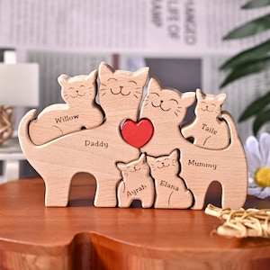 Könnte beinhalten: Holzskulptur einer Katzenfamilie mit den eingravierten Namen "Willow", "Daddy", "Ayrah", "Elana", "Tails" und "Mummy". Die Skulptur hat ein rotes Herz und ist aus hellem Holz gefertigt.