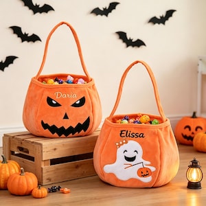 May include: Two orange plush Halloween candy buckets with handles. One bucket features a black jack-o'-lantern face and the name Daria. The other bucket features a white ghost holding a small pumpkin and the name Elissa. Both are filled with candy.