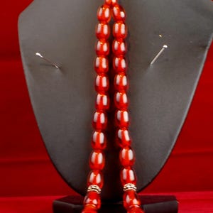 May include: A strand of translucent, reddish-orange prayer beads, displayed on a black stand against a red backdrop. The beads are oval-shaped and strung together. The top features a red tassel.