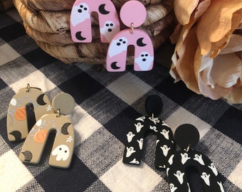 May include: Three pairs of Halloween-themed earrings. The first pair is pink with white ghosts and black crescent moons. The second pair is beige with orange pumpkins and black crescent moons. The third pair is black with white ghosts.