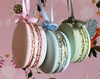 May include: Three pastel-colored macaron ornaments with gold glitter accents, hanging from pink, blue, and green ribbons.