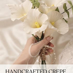 Könnte beinhalten: Ein Strauß aus handgefertigten Calla-Lilien aus weißem Krepppapier mit gelben Blütenmitten und grünen Papierblättern. Die Stiele sind mit Schnur umwickelt und mit kleinen weißen Perlen verziert. Text: EASY DIY GUIDE und HANDCRAFTED CREPE PAPER CALLA LILIES.