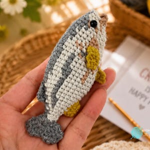 May include: A small, handmade crochet fish toy featuring a grey and white striped body, a dark grey tail, and small yellow fins. The fish has a single black bead eye and is crafted with a textured yarn stitch pattern, showcasing detailed needlework.