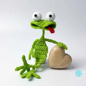May include: A handmade green crochet frog with large white eyes and a red tongue sticking out. The frog is posed next to a wooden heart. The frog's arms and legs are outstretched, giving it a playful appearance.