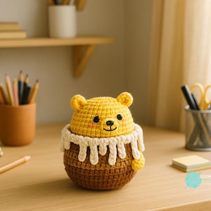 May include: A handmade crochet bear in a honey pot. The bear is yellow with black eyes and a smiling face. The honey pot is brown with a white, dripping honey design. The item is sitting on a wooden desk.