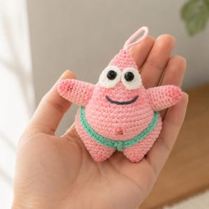 May include: A small, pink, crocheted starfish character with a pointed top, two arms, and two legs. It features large white eyes with black pupils, a stitched black smile, and a light green waistband. A small pink loop is attached to the top.