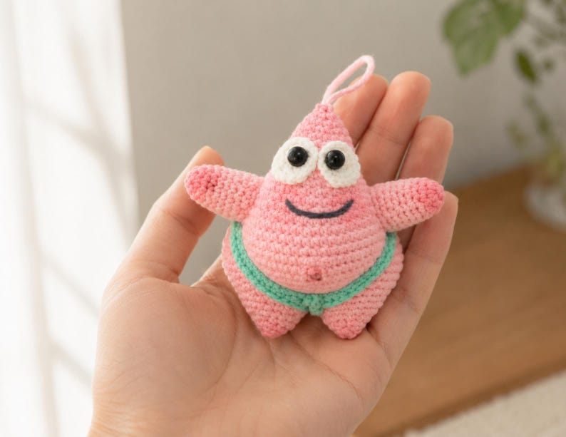 May include: A small, pink, crocheted starfish character with a pointed top, two arms, and two legs. It features large white eyes with black pupils, a stitched black smile, and a light green waistband. A small pink loop is attached to the top.