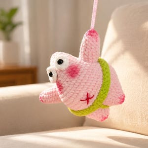 May include: A pink crocheted plush toy shaped like a starfish with a lime green waistband, large white eyes with black pupils, and a small red cross-stitch detail on its side. The figure hangs from a pink yarn loop attached to its top point.
