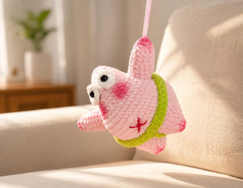May include: A pink crocheted plush toy shaped like a starfish with a lime green waistband, large white eyes with black pupils, and a small red cross-stitch detail on its side. The figure hangs from a pink yarn loop attached to its top point.