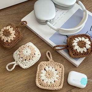 Puede incluir: Tres fundas cuadradas de ganchillo para auriculares inal&aacute;mbricos, cada una con un motivo floral central. Una funda es marr&oacute;n oscuro con centro crema, otra es crema con centro beige y otra es beige con centro crema. Cada funda incluye una peque&ntilde;a asa.