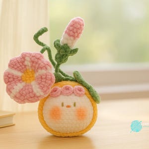 Puede incluir: Arreglo floral de ganchillo hecho a mano con una flor rosa y blanca, un capullo rosa y blanco y un personaje amarillo y blanco con mejillas rosadas. El arreglo está sobre una superficie de madera.