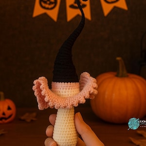 May include: A handmade crochet Halloween decoration featuring a witch's hat and collar. The hat is black, the collar is pink, and the body is off-white. A small pumpkin and a larger pumpkin are in the background, along with a lit lantern.