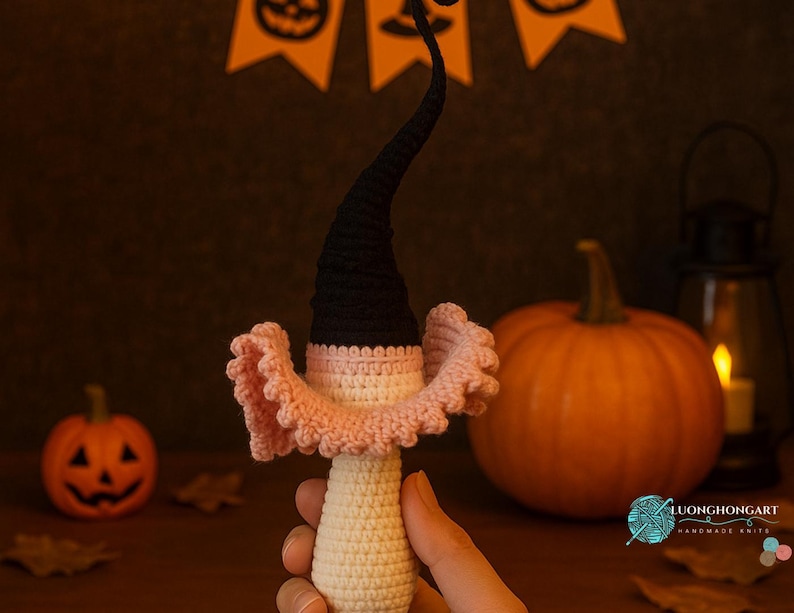 May include: A handmade crochet Halloween decoration featuring a witch's hat and collar. The hat is black, the collar is pink, and the body is off-white. A small pumpkin and a larger pumpkin are in the background, along with a lit lantern.