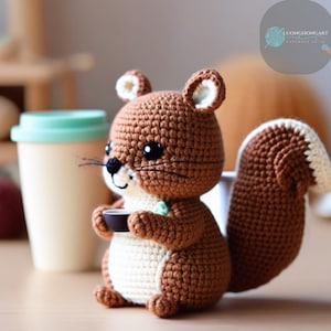 Peut inclure: Un écureuil en crochet marron et blanc tenant une petite tasse brune. L'écureuil est assis sur une surface en bois à côté d'une tasse verte et blanche.