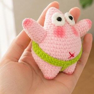 May include: A small, pink, crocheted starfish figure with a rounded body, two arms, and two legs. It features large white eyes with black pupils, pink blush on its cheeks, and a lime green waistband. A small red cross is stitched on its belly.