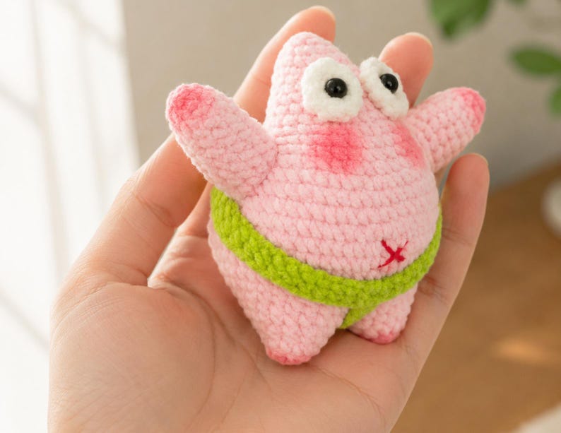 May include: A small, pink, crocheted starfish figure with a rounded body, two arms, and two legs. It features large white eyes with black pupils, pink blush on its cheeks, and a lime green waistband. A small red cross is stitched on its belly.