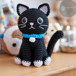 May include: Black crocheted cat amigurumi toy with white feet and a blue collar. The cat has black eyes and a pink nose.