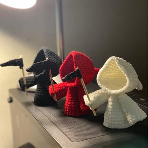 Pode incluir: Três figuras de crochê vestindo túnicas com capuz e segurando pequenas bandeiras. As figuras são pretas, vermelhas e brancas.