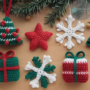 May include: A selection of handmade crochet Christmas decorations. The ornaments include a green Christmas tree, a red star, white and green snowflakes, and gift boxes in red and green. The items are displayed on a wooden surface.