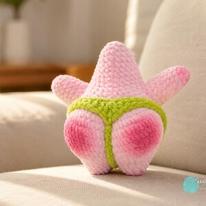 May include: A plush crochet toy shaped like a pink starfish with a textured yarn surface. The starfish features a bright lime green thong-style garment wrapped around its lower section. The overall design is soft, rounded, and handmade.