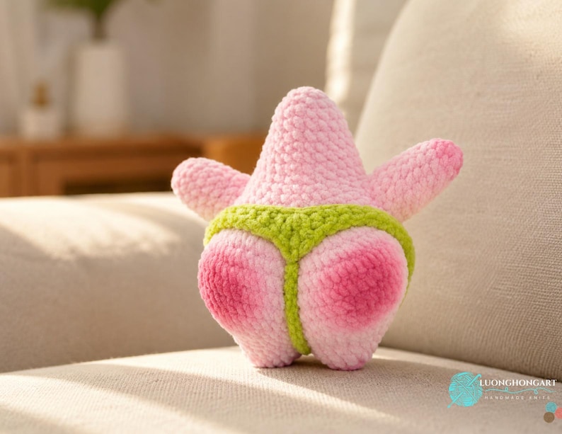 May include: A plush crochet toy shaped like a pink starfish with a textured yarn surface. The starfish features a bright lime green thong-style garment wrapped around its lower section. The overall design is soft, rounded, and handmade.