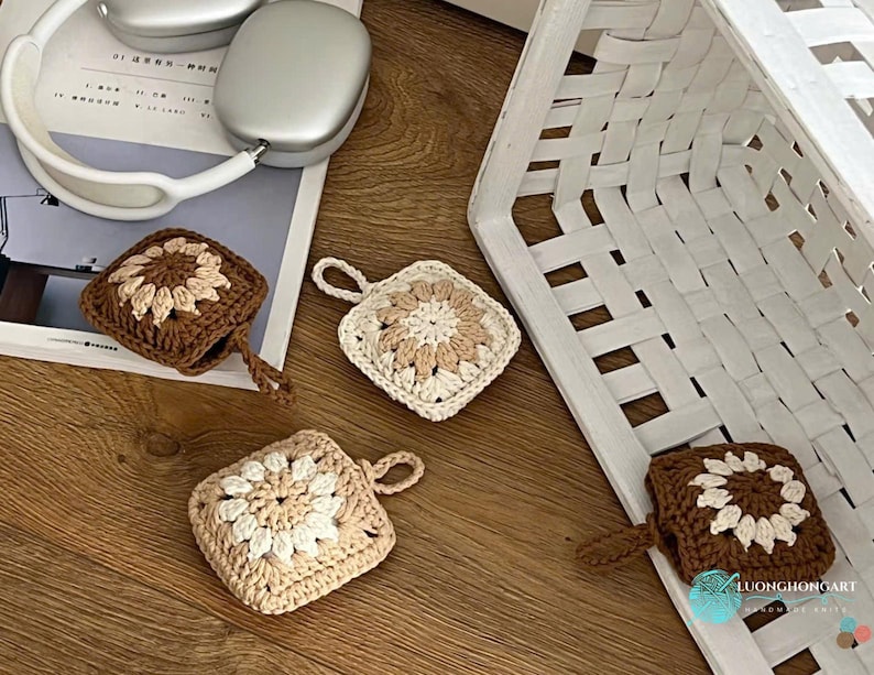 Puede incluir: Cuatro fundas cuadradas de ganchillo para auriculares inal&aacute;mbricos, con un motivo floral central en hilo crema y marr&oacute;n. Dos fundas son de color marr&oacute;n oscuro con centro crema, y dos son crema con centro marr&oacute;n claro, cada una con una peque&ntilde;a asa.
