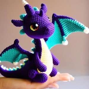 May include: A crocheted purple dragon with teal wings. The dragon has a white horn and yellow accents.