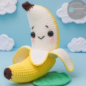 May include: A crocheted banana plush toy with a smiling face and a green leaf. The banana is yellow and white, and the leaf is green.
