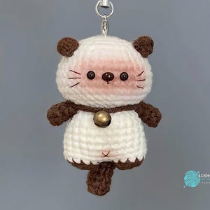 May include: A handmade crochet keychain in the shape of a cat. The cat has a white body, brown ears, and a brown tail. It has a pink blush on its cheeks, brown eyes, and whiskers. A small gold bell hangs from its neck.