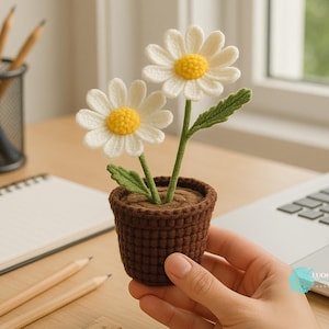 May include: A handmade crochet daisy flower arrangement in a brown pot. The daisies have white petals and yellow centers, with green stems and leaves. The pot is held by a person.