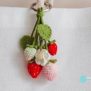 Op de afbeelding: Een handgemaakte gehaakte aardbei sleutelhanger met een zilveren metalen ring. De sleutelhanger heeft rode, roze en groene aardbeien, een witte bloem en groene bladeren. De sleutelhanger is bevestigd aan een witte canvas tas.