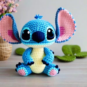 Op de afbeelding: Een blauw en roze gehaakt Stitch knuffel met grote ogen en een zwarte neus. Het speelgoed zit op een houten oppervlak.