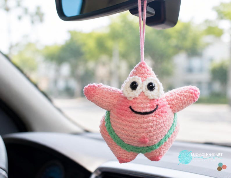 May include: A pink crochet starfish ornament with a smiling face, black eyes, and a light green waistband. The character has a pointed top and is suspended by a pink string. A small logo with the text LUONGHONGART is visible in the bottom corner.