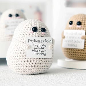 May include: Two crocheted potato plushies, one white and one brown, each holding a sign with a positive message. The white potato says "Positive potato I may be a tiny potato but I believe in you Go do your thing!" The brown potato says "was a potato rolead".