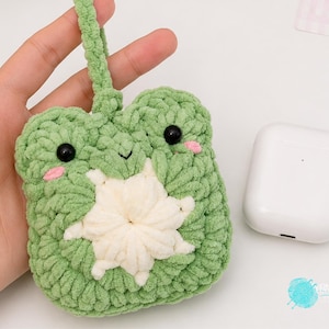 Puede incluir: Una funda de ganchillo de color verde claro con forma de cara de rana, ojos de cuentas negras, mejillas bordadas en rosa y un detalle central en forma de estrella blanca. La funda incluye un lazo de ganchillo verde a juego en la parte superior.