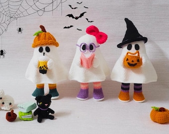 May include: Crocheted Halloween ghosts in white with orange, black, and purple accents. One ghost wears a pumpkin hat and holds a drink, another wears glasses and reads a book, and the third holds a jack-o'-lantern bag.