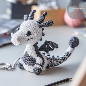 Könnte beinhalten: Ein schwarz-weißer gehäkelter Drachen-Schlüsselanhänger mit einem silbernen Schlüsselring. Der Drache hat einen kleinen, eingerollten Schwanz und ein Paar Flügel.