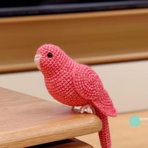 May include: A handmade, crochet bird in shades of pink, with a white beak and black eyes. The bird is perched on a wooden surface. The bird's tail is extended. The bird is a decorative item.