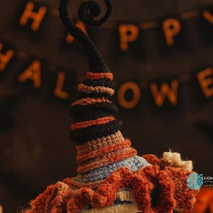 May include: A handmade crochet witch hat in black, orange, and blue, with a curly top. The hat is decorated with orange and brown yarn. The background includes a "HAPPY HALLOWEEN" banner and a lit candle.