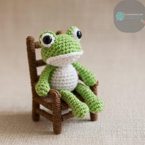 Puede incluir: Una rana de crochet verde sentada en una silla de madera marrón. La rana tiene detalles de crochet blanco y ojos de botón negros.