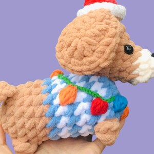 May include: A handmade crochet dog toy in shades of tan, white, and blue, wearing a Santa hat and a festive collar with colorful lights. The dog is held in a hand against a purple background. A charming holiday decoration.