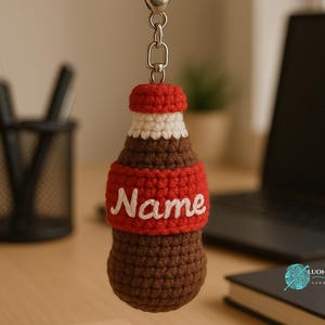 May include: Crocheted keychain shaped like a soda bottle. The bottle is brown with a red band featuring the name "Name" in white script. The top is red and white, attached to a silver chain and keyring. The keychain is handmade.