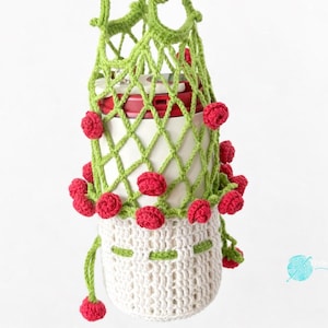 May include: A handcrafted crochet plant holder in white, green, and red. The plant holder features a white pot with green trim and a green net-like design. Red crochet flowers are attached to the green net. The plant hanger is suspended against a white backdrop.