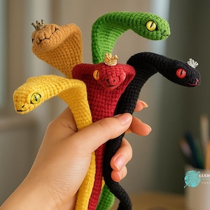 Può includere: Una mano tiene cinque giocattoli a forma di serpente all'uncinetto in giallo, arancione, verde, rosso e nero. Ogni serpente indossa una corona e ha occhi ricamati. I serpenti di filato hanno un aspetto stravagante e fatto a mano.