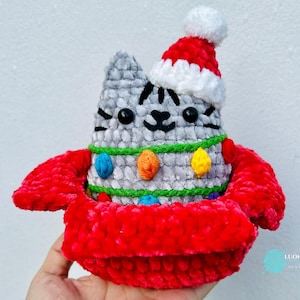 Op de afbeelding: Handgemaakte gehaakte kattenfiguur met een kerstmuts en een rode kersttrui. De grijze kat heeft zwarte strepen en is versierd met kleurrijke lichtjes. Een feestelijke handgemaakte decoratie voor de feestdagen.