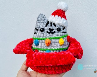 May include: A handmade crochet cat figurine wearing a Santa hat and a red Christmas sweater. The gray cat has black stripes and is decorated with colorful lights. The figurine is a festive holiday decoration.