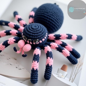 Puede incluir: Un juguete de araña de crochet con un cuerpo negro y patas rosas. La araña tiene ojos grandes y negros y una boca rosa.