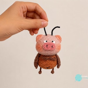 May include: A crocheted pig figurine with a light pink head, a darker pink snout, and a brown striped body. The character features two black antennae on top of its head, small black eyes, and two small arms and legs. The text LUONGHONGART is visible.