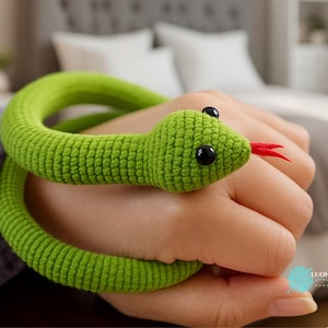 Puede incluir: Un juguete de serpiente de ganchillo verde con ojos negros y una lengua roja. La serpiente está enrollada alrededor de una mano. El juguete está hecho de un hilo suave y texturizado. La cabeza de la serpiente está detallada con ojos negros y una lengua roja bifurcada.